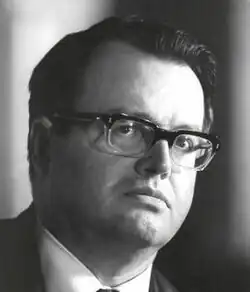 Three-quarter profile publicity shot of a clean-shaven man with serious expression, spectacles, and black hair combed back.
