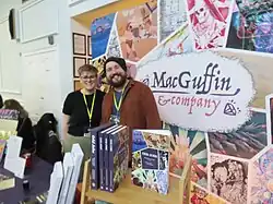 Jonathan Sims and his spouse Sasha Sienna stand inside a large Macguffin & Co booth at the Dungeons and Flagons Christmas stall, in front of a sign showing the company logo, game art, and advertising "Odd Jobs".
