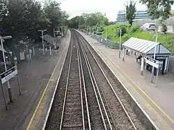 The view back from the footbridge. The pair of 3rd-rail-electrified tracks curve to the left between the 2 platforms. the left one has a sign with the number 2, stairs & a help-point. They both have a shelter, a ticket machine, display boards & lamp-posts. The trees on the left are darker in colour than the trees on the right. There is a silver car on the right & a modern office building.