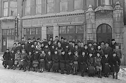 The image shows a large group of people lined up in three rows in the foreground.