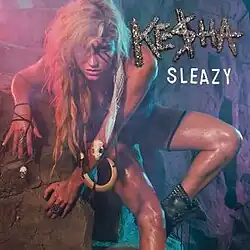 A blond-haired woman rests on a rock wall. She wears tribal clothes: a dark suit, boots and bone ornaments. She has three strips painted across her left eye and a skull, and a long white strip, throughout her left arm. Over her left shoulder the words "Ke$ha" and "Sleazy" are written.