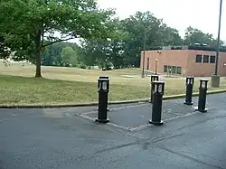 Color photograph of a memorial (six posts with lights set around a rectangular demarcation) with grass, trees, and a building in the background.