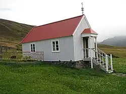 A small white church with red roof. It is on uneven ground, and a short set of stairs leads to the door.