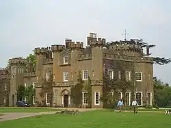 A battlemented stone building with two people, a dog, and a car in front.