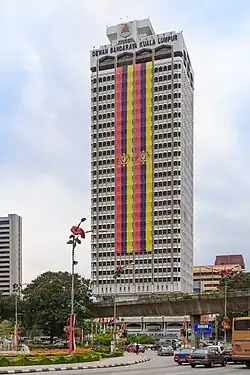 Tall white building with two long flags of vertical red, blue, and yellow stripes and the Malaysian coat of arms hanging down most of the building's length
