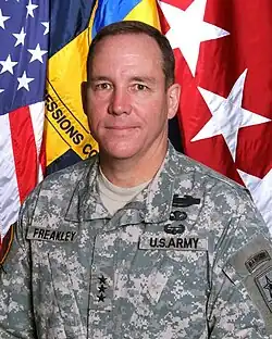 Portrait photo of Caucasian male wearing an army combat uniform with the rank of lieutenant general, Combat Infantry Badge, Basic Parachutist Badge, Air Assault Badge, and a Ranger tab standing in front of the U.S. flag, a command flag, and a lieutenant general flag.