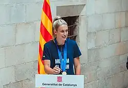 Alexia Putellas stands behind a podium at the Palau de la Generalitat de Catalunya in Barcelona, Spain. She is wearing her Champions League winners' medal over a t-shirt that features Barcelona Femení's victory slogan "Movem el mon" ("We move the world" in Catalan).
