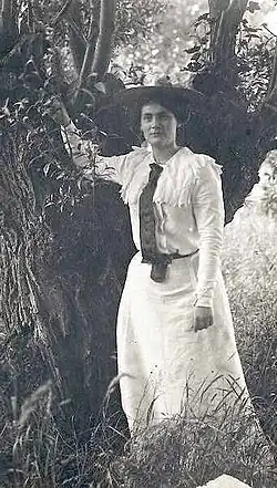 young woman in full-length frock and top coat leaning on a tree