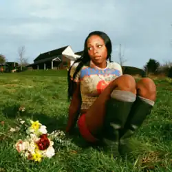 Chinouriri sitting in the grass and looking to the left, next to a flower bouquet. She is in front of a house and rural landscape on a cloudy day.