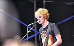 Byrne is singing into a microphone on its stand. He has a guitar strap but the instrument is out of shot. Behind him are blue beams from lights. He wears a David Bowie/Aladdin Sane t-shirt. He has curly gold-red hair with his eyes closed and veins popping in his neck.