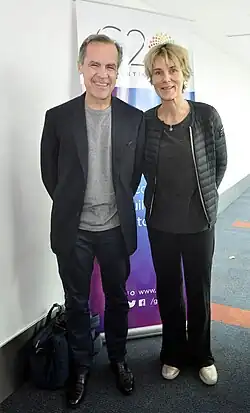 Mark Carney stands in front of a vertical banner for the 2018 G20 Summit with his wife, Diana Carney. He wears a dark blazer, pants, and a grey shirt, while she wears a dark jacket, shirt, and pants.