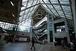 A large glass-roofed lobby with elevators, stairs, and escalators leading to an adjacent parking garage