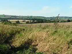 In the foreground, a wide ditch with a grassy bank. In the distance, fields bordered by hedges and trees. In the background, low hills covered with meadows and copses.