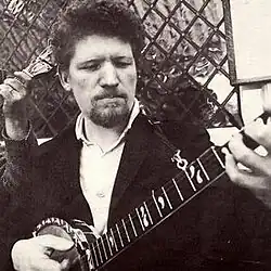 Black and white picture of a man (with focus on torso, not a full length picture). He has goatee (beard) and curly hair, and is holding a banjo He is looking at the banjo with concentration. He is wearing a shirt and coat.