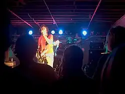 A band is performing on stage with three members visible. A man is at front left, he is singing into a microphone, which is held in his left hand. He has a guitar slung over his shoulder. The drummer sits behind a kit in the centre. A bearded man is at the right but is blurred. All three artists are partly obscured by audience members. There are stage lights behind and above the artists. Musical speakers are partly visible at right.