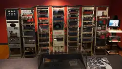 A series of seven tall metal racks filled with electronic equipment standing in front of a brick wall. Signs above each rack describe the functions carried out by the electronics they contain. Three visitors read from information stands to the left of the image