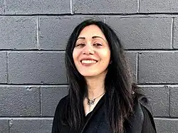 Woman with black hair, a black coat, and a silver necklace smiling into the camera, with a gray cinderblock wall in the background