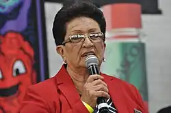 Picture of woman with sort black hair and glasses, in red jacket, holding microphone