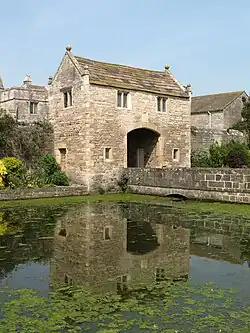 A two-storey stone building with a vehicular arch through the centre. It has a stone roof and the gables at each end have stone ball finials. In front of the gatehouse is a stone bridge across a wide moat that has water plants growing on it.