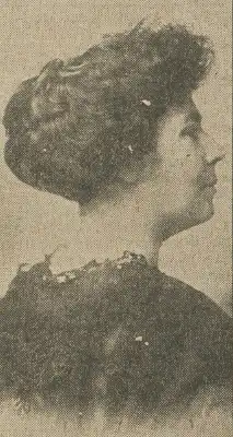 A white woman photographed in profile; her dark hair is in an updo; she is wearing dark garment