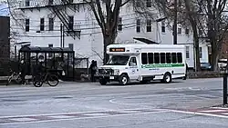A white and green minibus stops to pick up passengers, including two bicyclists.