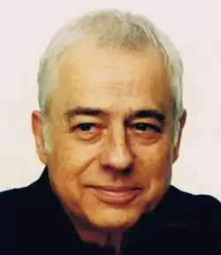 Colour publicity shot of a half-smiling, clean-shaven man with dark eyes and eyebrows, strong features, and short white hair brushed forward