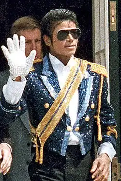 Jackson is wearing sun glasses and a black sequinned jacket with gold shoulders and a golden sash running diagonally across his chest. His right hand is raised in a wave; he is wearing a white glove on it. His skin is dark