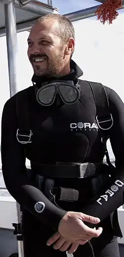 Mike Rutzen, wearing a black wetsuit and scuba gear, stands on a boat smiling as he prepares to dive with a Great White Shark for a nature documentary.