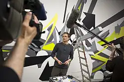 Mirko Reisser wearing a dark long-sleeved t-shirt and dark green striped pants, standing next to a ladder in front of his artwork walltaping, looking right, with several small items in his hands, camera crew in the foreground
