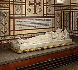 This image shows the monument to Baroness Coleridge (d.1879) by Frederick Thorpe at Ottery St Mary, Devon, exemplifying the Victorian Gothic Revival's return to medieval monument forms. The white marble effigy depicts the Baroness lying in peaceful repose on a low tomb chest, her hands folded in prayer in the traditional medieval manner. At her feet lies a small dog, following the medieval convention of placing symbolic animals as footrests. The monument is set within an elaborate polychromatic mosaic surround featuring geometric patterns, crosses, and floral motifs executed in cream, brown, red, and blue tesserae, characteristic of Victorian decorative arts. Above the effigy, a lengthy inscription panel records details of the memorial's dedication. The tomb chest itself displays colourful mosaic decoration that complements the wall treatment. This monument demonstrates how Victorian designers combined authentic medieval effigy poses with contemporary decorative techniques, creating works that were both historically inspired and distinctly of their own period. The rich colour scheme reflects the Victorian fascination with medieval polychromy and Byzantine artistic traditions.
