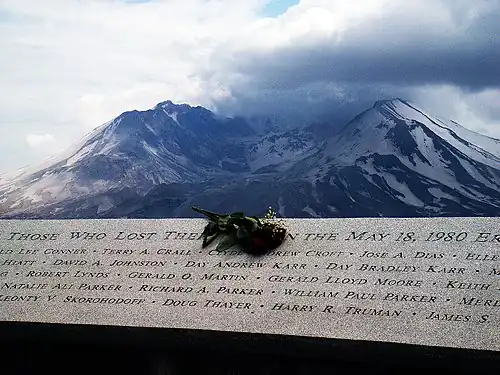A plaque with the carved names of the eruption's victims appears, with a bouquet of flowers sitting on its center. In the background, Mount St. Helens can be seen.