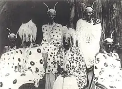 A group of Rwandan royal family members sit and stand in front of a hut, adorned in their royal regalia.