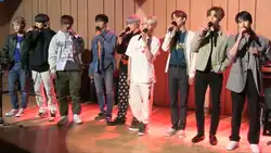 Nine men wearing a mix of casual and stylish outfits, with variations including jackets, hoodies, and t-shirts, standing on a wooden stage, each holding a microphone and singing with some having dyed hair in shades of silver and blue.