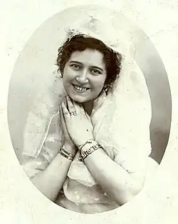 A young white woman with dark curls, smiling, hands together under her chin; she is wearing light-colored lacy clothes including a headpiece, and several bracelets