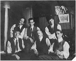 A black and white photograph of six young-to-middle-aged men, all wearing white shirts, dark ties and waistcoats.