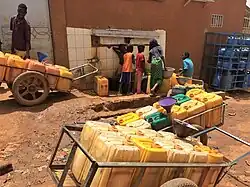 A family receives water from a standpipe. Some metal pushcarts, each containing ten jugs, surround the standpipe.