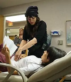 An exam room set, getting ready for a take. Nikole, center, with long dark hair, bangs, wearing black shirt, black pants, black hat, headphones and a utility belt with a walkie clipped on her hip stands over actress Patti Harrison who is laying on an exam table in a white shirt and red sweat pants holding a script in her left hand. They look at each other as Nikole gives direction. Other crew members hug the perimeter of the room.