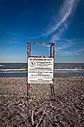 A white sign attached to a wooden structure indicating to patrons on a beach. "No Lifeguard On Duty, Call 911 in case of emergency"