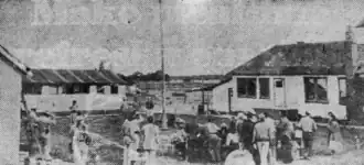 A black-and-white newspaper photograph published in the August 12, 1954 edition of the Nome Nugget. A crowd of men, dressed in pants and shirts and belts, stand in a courtyard outside a single-story schoolhouse. A pole (possibly a flagpole?) appears to be in the middle. At the edges on the left appear to be men in hats (possibly law enforcement?) and on the edge at the right is what appears to be a woman in a dress.