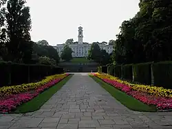A white building topped by a tower, at the end of a flowery driveway.