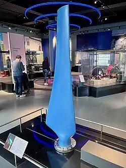 A blue turbine blade about 2m high, mounted vertically with a metal adaptor bolting it to a plinth, in an exhibition gallery of the National Museum of Scotland