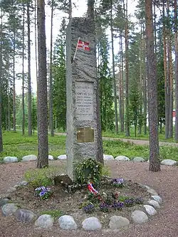 A stone monument in a forest clearing, adorned with a plaque and small Norwegian flag