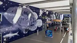 Photograph of an interior space with one wall covered by blue and white tiles, that looks like a pixelated image of various fish from afar