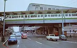 A white-and-green monorail train hangs over a city street with several cars.