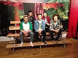 Waxwork of the members of One Direction created by Madame Tussauds. They sit on a bench against a cloth background of a tree with a red trunk. In the front row, from left to right: Niall Horan, Louis Tomlinson, and Harry Styles. In the back row, from left to right: Zayn Malik and Liam Payne.