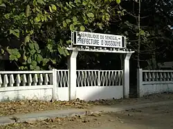 A white gate with a sign reading "Republique du Senegal: Prefecture d'Oussouye"