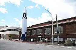 An exterior photo of an upright rocket standing outside of a brick building