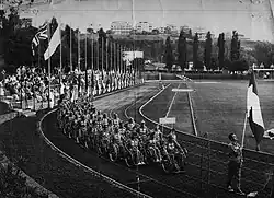 A black and white photo of a large track and field stadium with spectator stands and flags flying on the left hand side and trees in the background. A large procession of people in wheelchairs goes round the track with a sign being held at the front reading "Italia". The team is led by a walking man carrying the Italian flag.