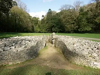 Front view of cairn, its boulders retained by a short, coursed, dry-stone wall that forms a bell-shaped courtyard at its entrance. The cromlech is set in flat ground of short grass. Trees are mainly in leaf to its rear.