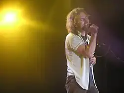 A male singer, Eddie Vedder, onstage and singing into a vocal microphone. He has an emotional look on his face as he sings.